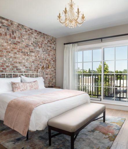 A king bed with white blanket and pink accents with a bench at the foot of the bed, Large glass doors overlook greenery