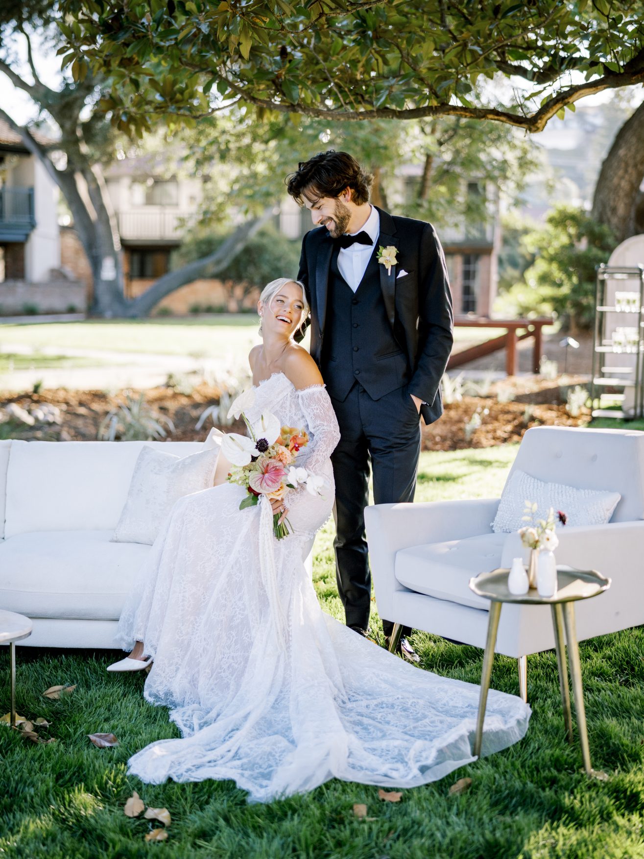 Bride in white sitting on white sofa looking at groom in black in a garden.