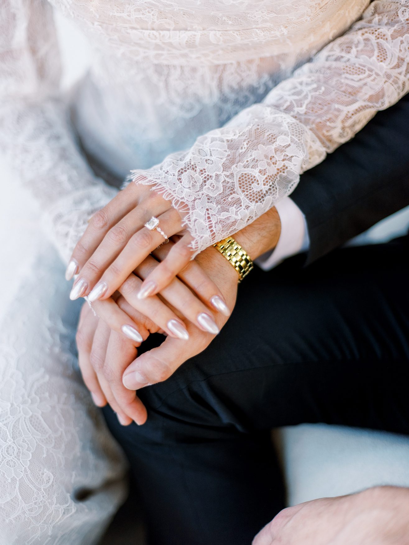 Bride in white long lace sleeved dress with hands on top of groom in black with a gold watch on his hand.