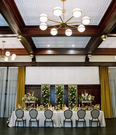 Formal dining table wiht 12 chairs. Candles and flower arrangements on top of the table and behind it on the wall. 